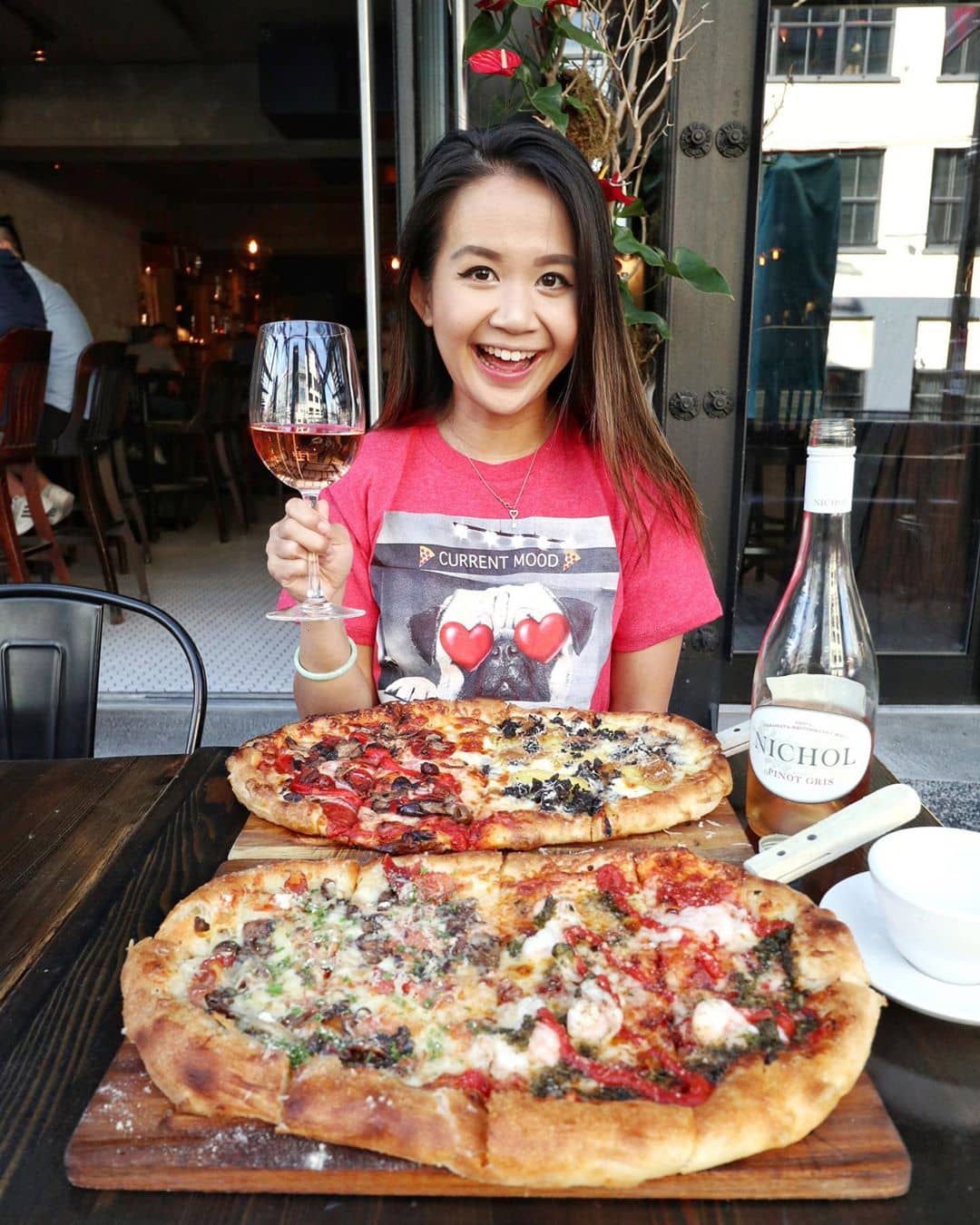 girl sitting infront of two pizzas holding a glass of wine
