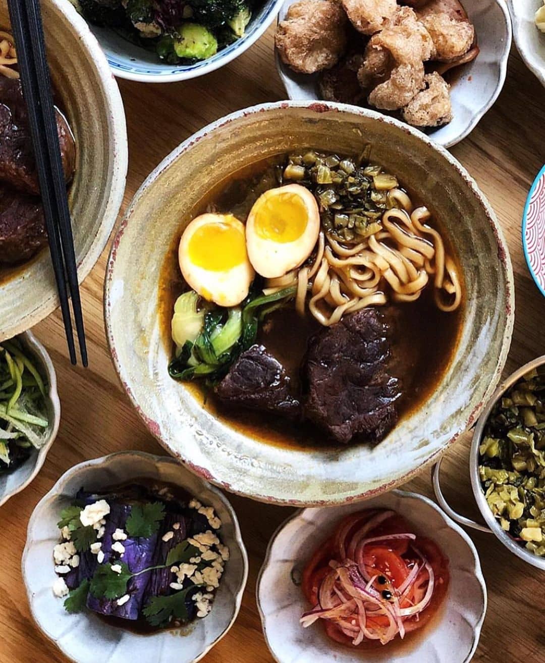 big clay bowl of taiwanese beef noodles with one egg sliced in half and side dished around it