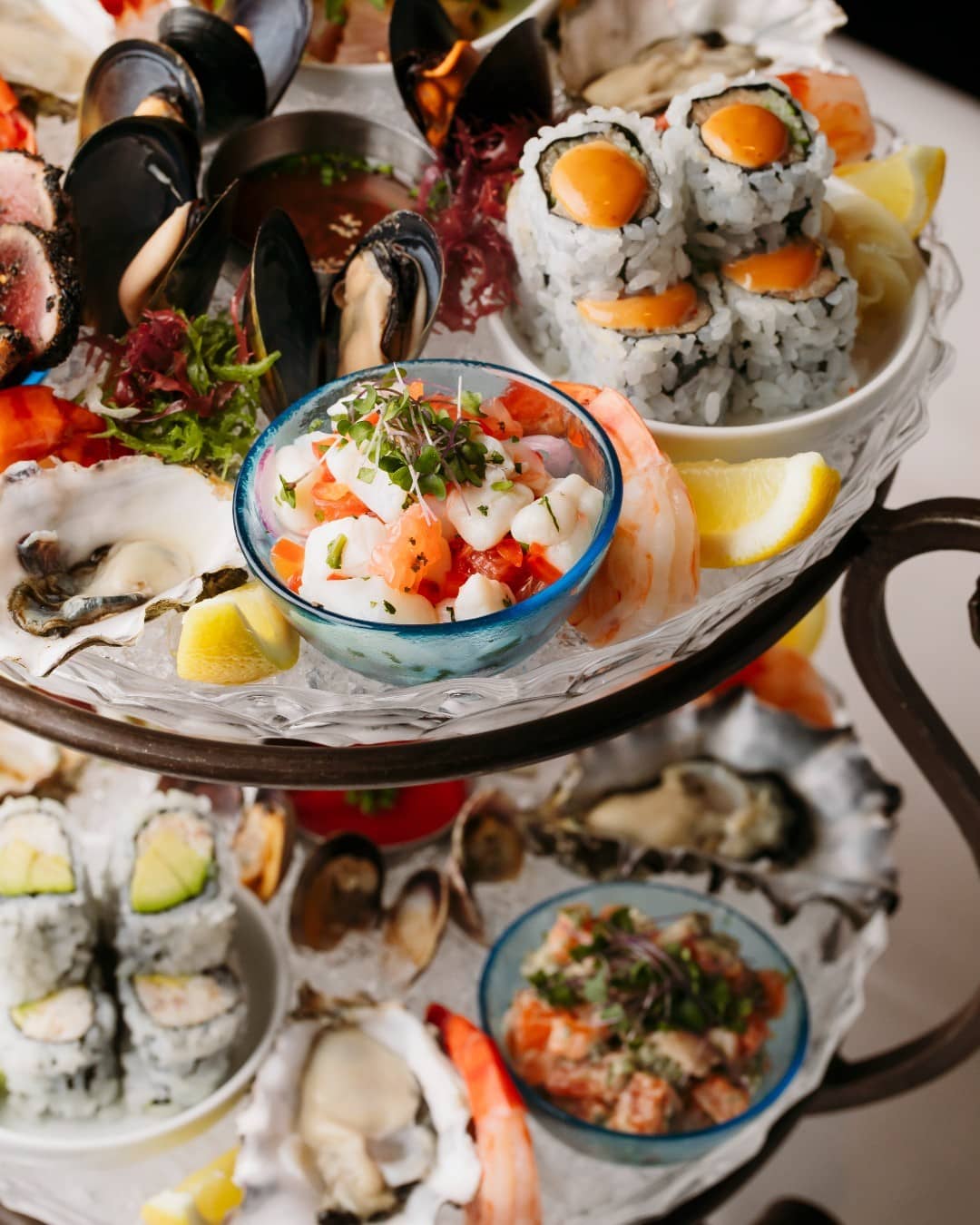 A photo of a three-tiered seafood tower on a white tablecloth at Blue Water Cafe in Vancouver, Canada. The tower features oysters, prawns, lobster, mussels, clams, and other seafood items on ice. (Photo Credit: Blue Water Cafe via Noms Magazine Ltd.)