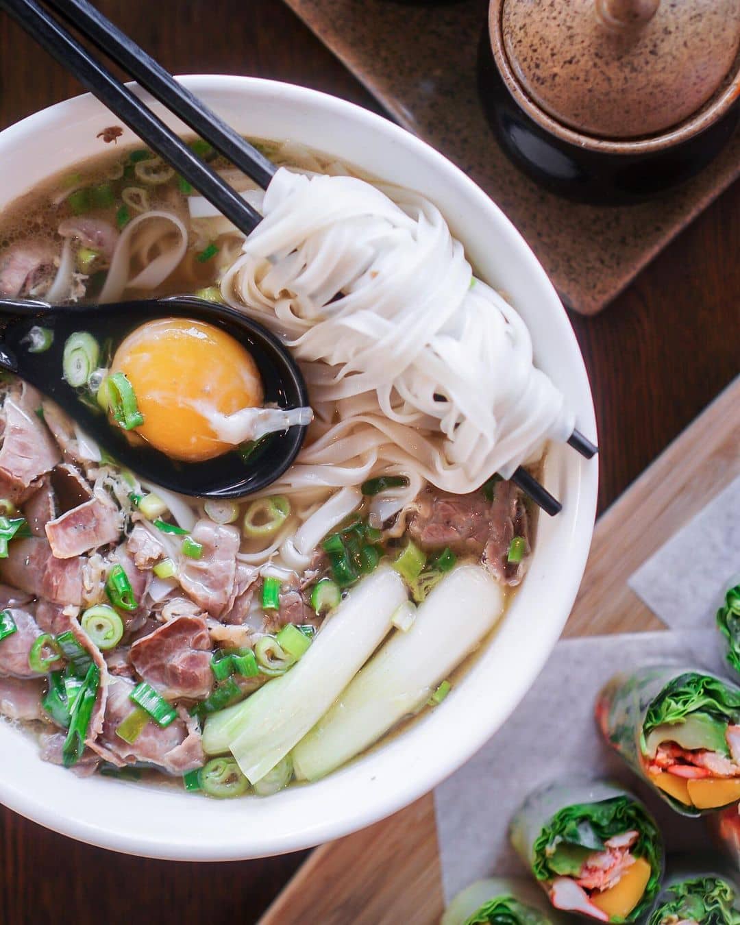 bowl of pho with egg yolk in spoon and salad rolls on wooden board best west vancouver restaurants - wooden fish